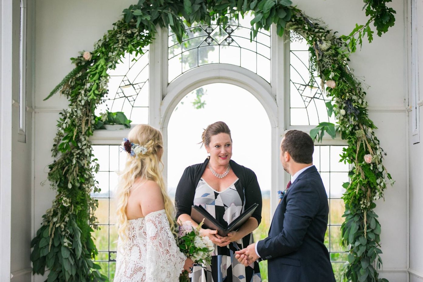 A bride and groom stand in front of the chapel window with their marriage celebrant.